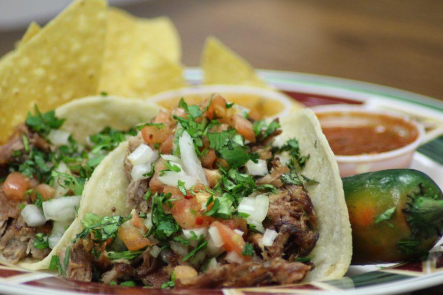 Two carefully prepared carnitas tacos de Las Palmas  garnished with cilantro and onion sit on a plate with chips, two types of homemade salsa and a jalapeno. 