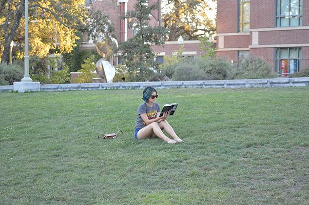 Kai Schade rests on the lawn in front of Doyle Library enjoying a book and sporting her blue-green hair.