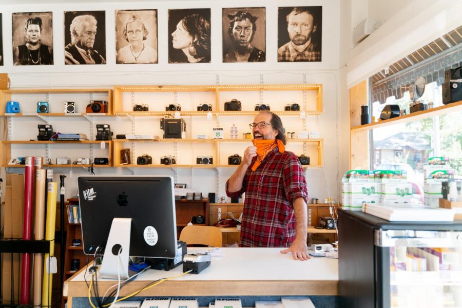 A man wearing a mask behind a cashier desk surrounded by tin-type portraits