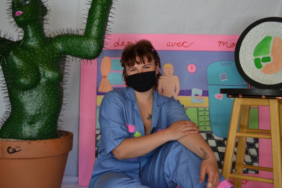 Sydney Gittins, a girl with reddish brown hair and bangs wearing a painted blue jumpsuit, sits in between a few of her art pieces (a cactus woman statue on the left, behind her is a drawn art piece set in a kitchen and on the right is a painted stool).