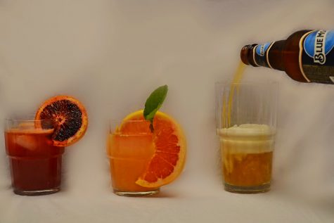 Three drinks in a row on a table. The left-most drink is a blood orange kahlua cocktail, the middle is a greyhound cocktail and the last is a beer which is currently being poured into the glass from off screen.