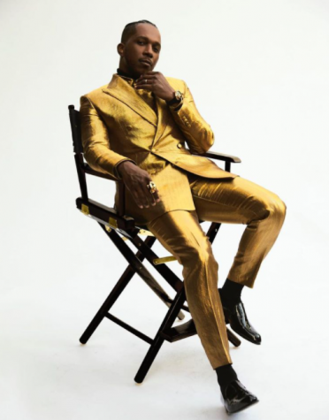 Leslie Odom Jr. sits on a dark director's chair wearing a gold Brioni suit.