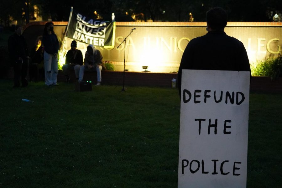 A photo of a person carrying a sign that says "Defund the police." Behind them is a group of people holding up a "Black Lives Matter" flag.