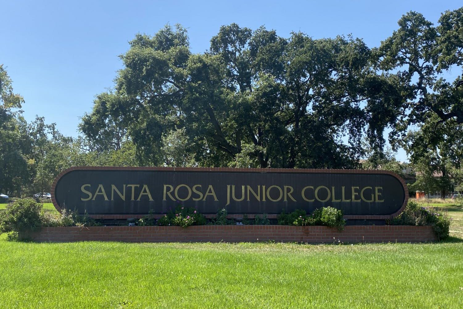 A sign for Santa Rosa Junior College stands on a green lawn backed by large oak trees and a blue sky.