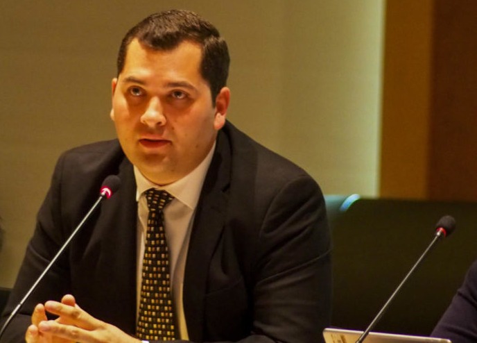 A man in a suit, Jordan Burns, sits at a desk and speaks into a small microphone.