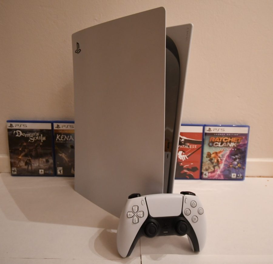 A silver PS5 sits on top of a white table with four PS5 games leaning against a white wall behind it. A primarily white controller with black accents sits in front of the PS5.