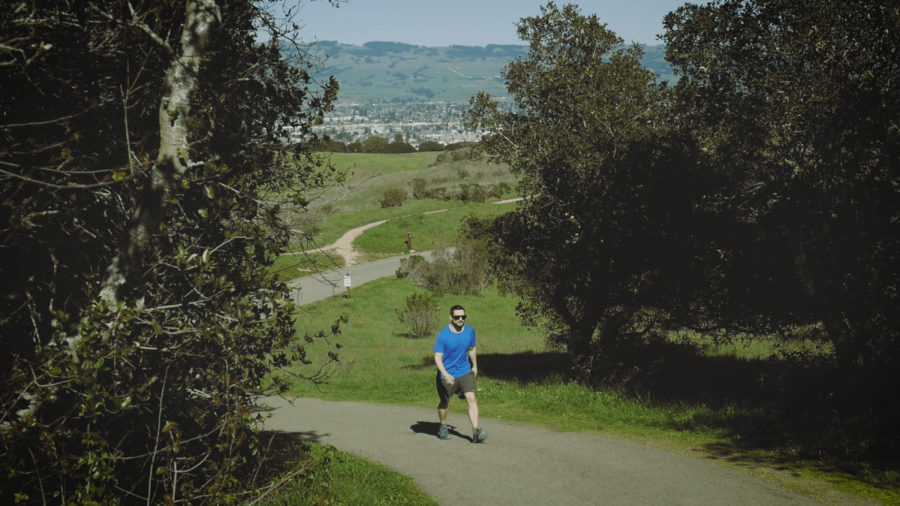 A masculine presenting person walks up a dirt path between trees on a clear day at Helen Putnam Park.