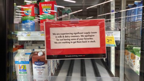 An empty shelf behind glass next to milk in a grocery store with a sign that says "We are experiencing significant supply issues in milk & dairy creamers. We apologize for not having some of your favorites. We are working to get them back in stock."