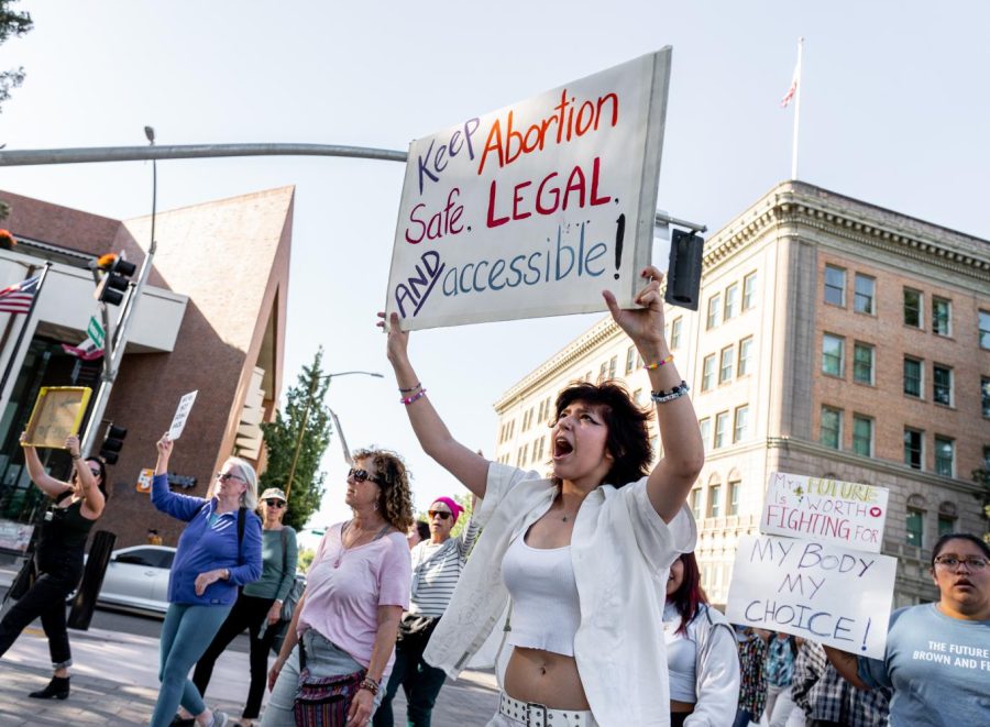 Many people marching in a protest. One person is in the foreground with a white sign that says "Keep abortion safe. Legal. and Accessible!"