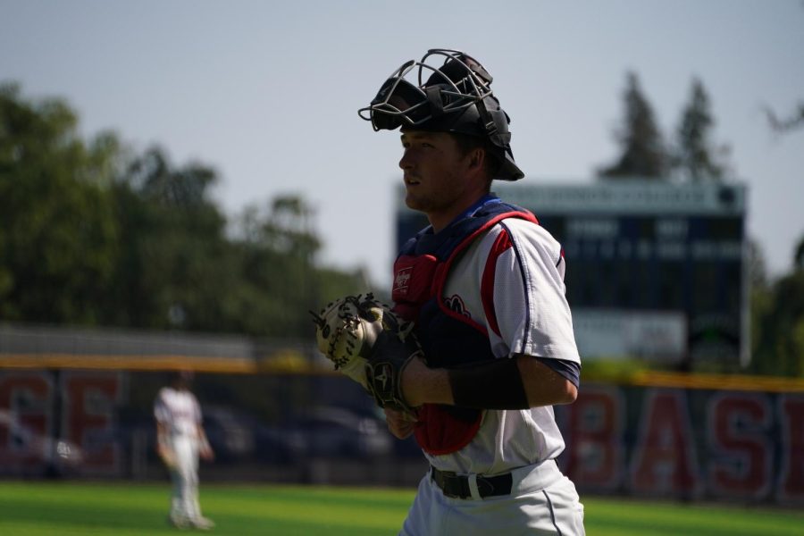 A baseball catcher in uniform in front of a blurry baseball scoreboard.
