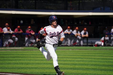 A baseball player runs toward the camera on a grassy baseball field with the dugout blurred in the background.