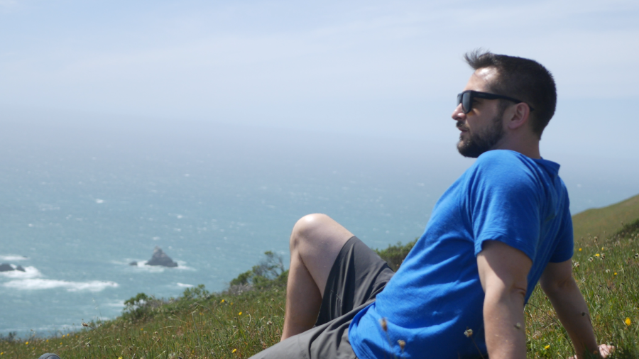 Michael Combs sits on a slanted grassy hill overlooking the ocean on a sunny day.
