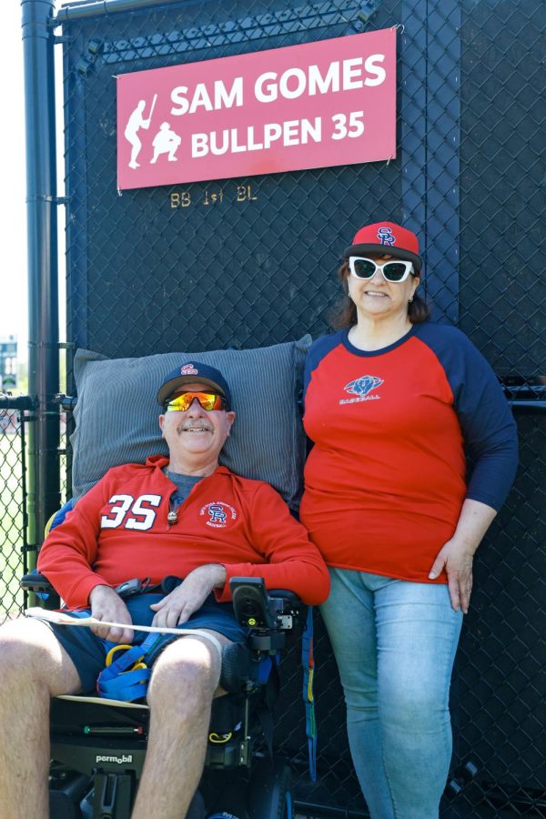 Sam Gomes in his wheelchair wearing a bright red SRJC baseball jacket, sunglasses and a blue SRJC hat, next to his wife, also wearing a bright red SRJC baseball shirt, sunglasses, and a red SRJC hat. They are in front of the bullpen wall, which says SAM GOMES BULLPEN on a red sign with white letters.
