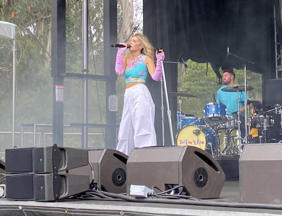 A girl wearing white pants and a teal shirt with pink arm warmers sings into the mic
